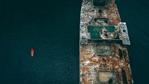 Aerial view looking down on a weather streaked concrete ship hulk, dwarfing the red kayak paddling next to it.