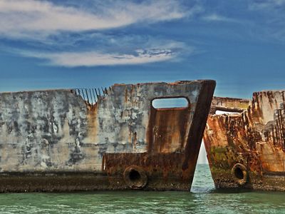 Two weather streaked concrete ship hulks sit nose to nose to create a breakwater protecting a calm, shallow recreation area.