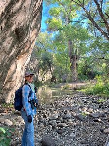 A woman wearing hiking gear and binoculars stands in front of a creek and vegetation, smiling at the camera.
