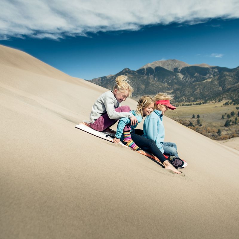Three kids surf down sand dune.