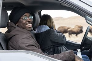 A young man in glasses smiles, sitting in a car, as he watches bison in the nearby prairie.