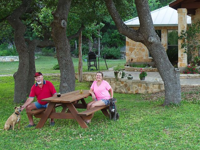 A man and woman sit with two dogs at a picnic table in front of a stone house.