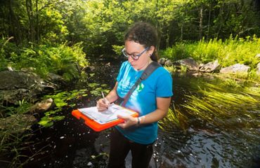 Laura Marx stands in a stream surrounded by greenery, holding a clipboard and filling out the form on it.