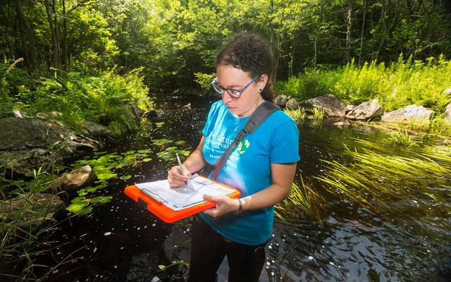 Laura Marx stands in a stream surrounded by greenery, holding a clipboard and filling out the form on it. 