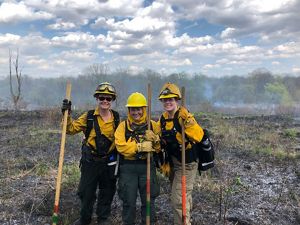 Three staff members stand in front of a controlled burn at Ives Road Fen. They're wearing protective gear while standing in the remains of the fire location. 