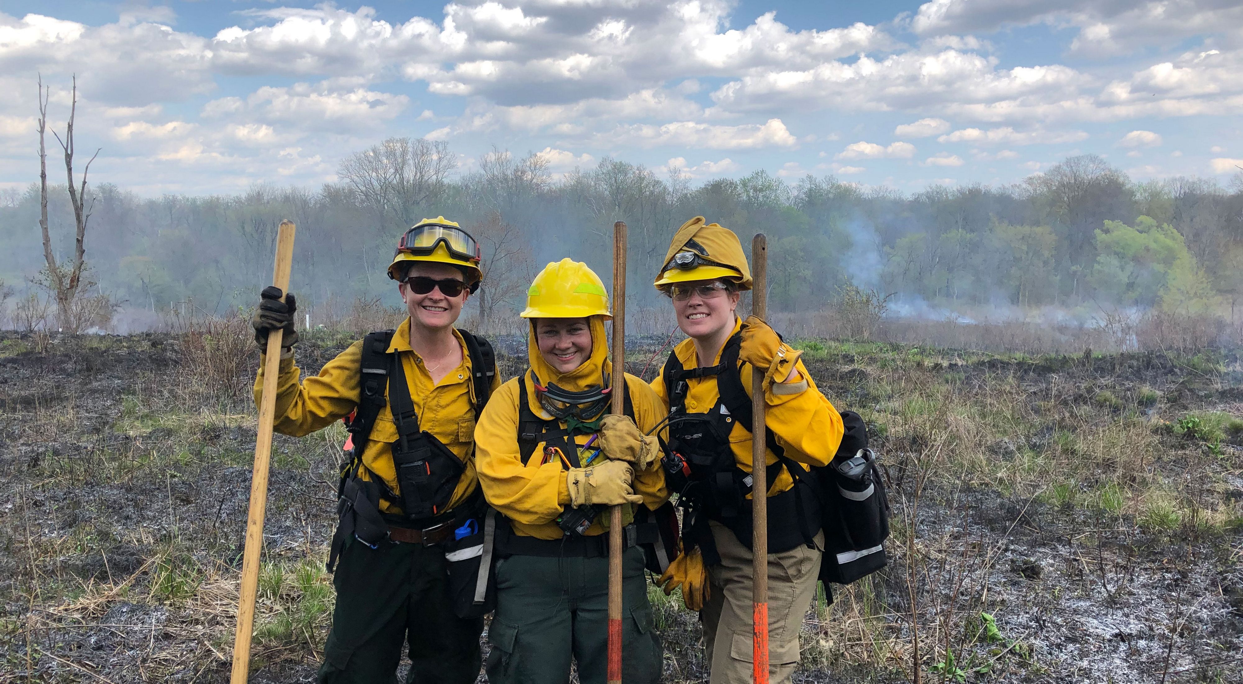 Three women holding shovels and wearing hard hats and burn gear at a prescribed burn in a field.