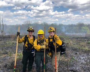 Three women holding shovels and wearing hard hats and burn gear at a prescribed burn in a field.