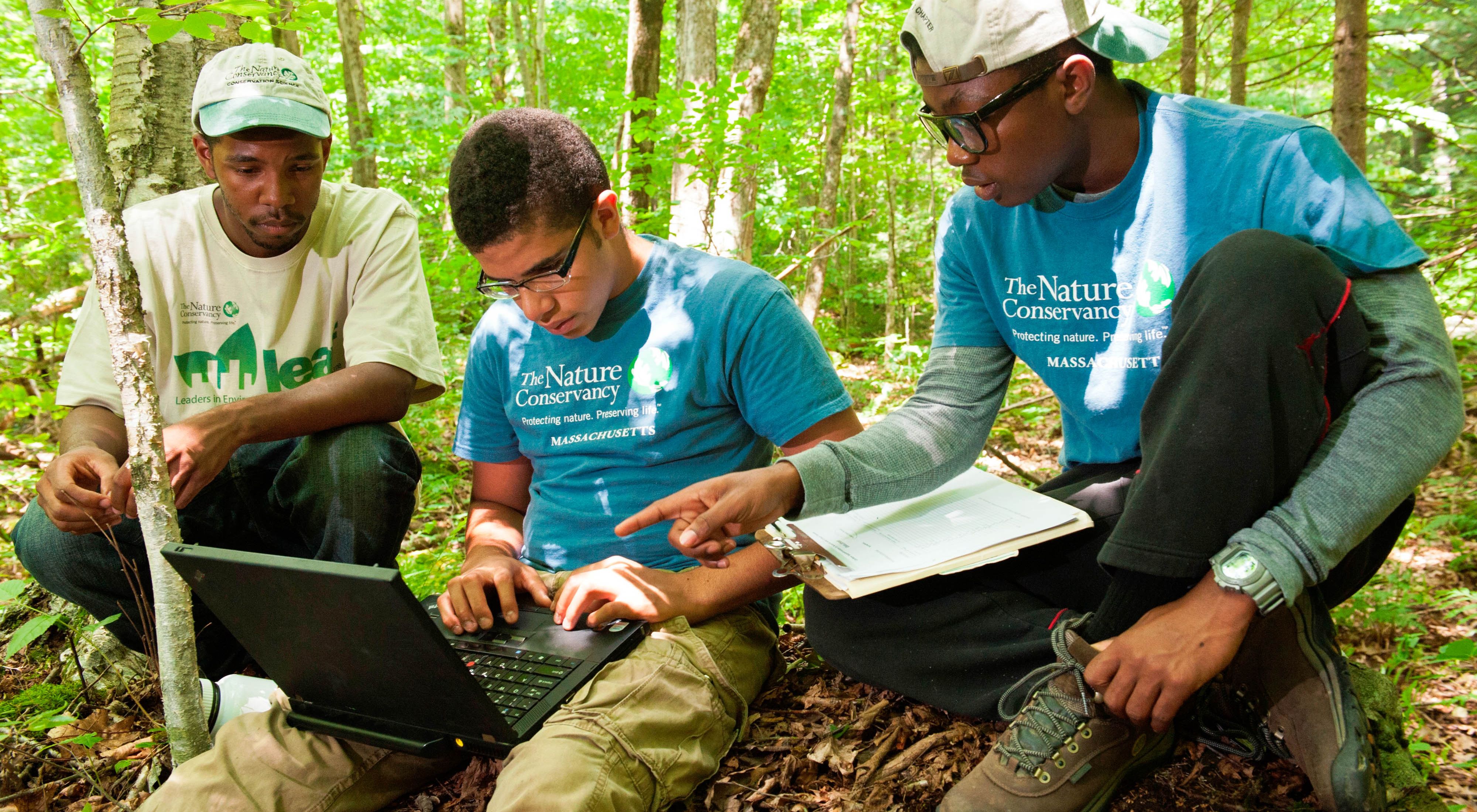 Three young people work on a laptop computer in a forest.