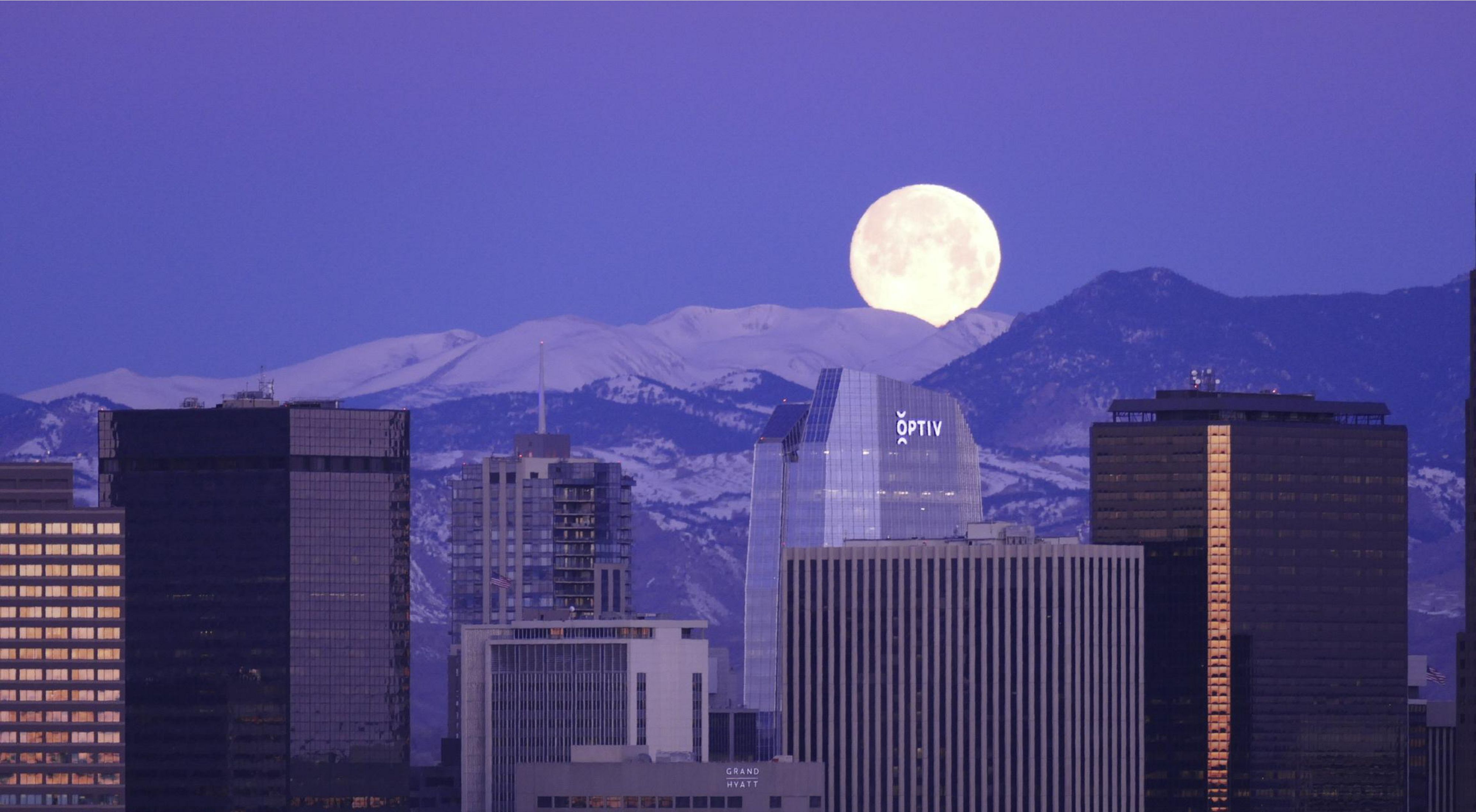 A huge moon rising over snow-capped mountains behind the Denver skyline.