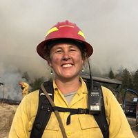 Women in yellow fire gear & red hardhat stands by prescribed burn.