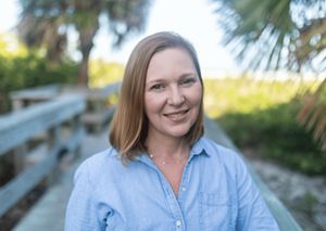 A woman stands near coastal dunes and palms; she has short red hair and blue-green eyes. 