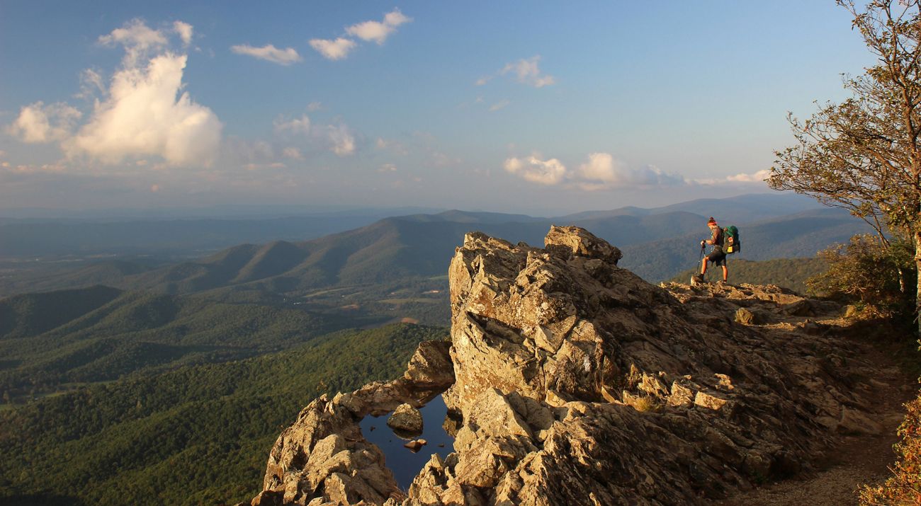 A man wearing a green backpack stands on a rocky overlook above a green forested valley. The setting sun reflects in a pool of water that has collected in the rocks.