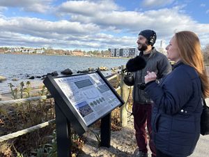 Podcast Producer Jay Feinstein, holding a mic and wearing headphones, stands with CZM's Julia Knisel in front of a sign about salt marshes, looking out at Collins Cove
