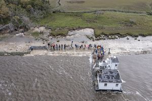 Several people stand along a shoreline in front of a white barge.