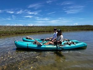 Two people sit side by side in teal blue kayaks in a shallow coastal area.