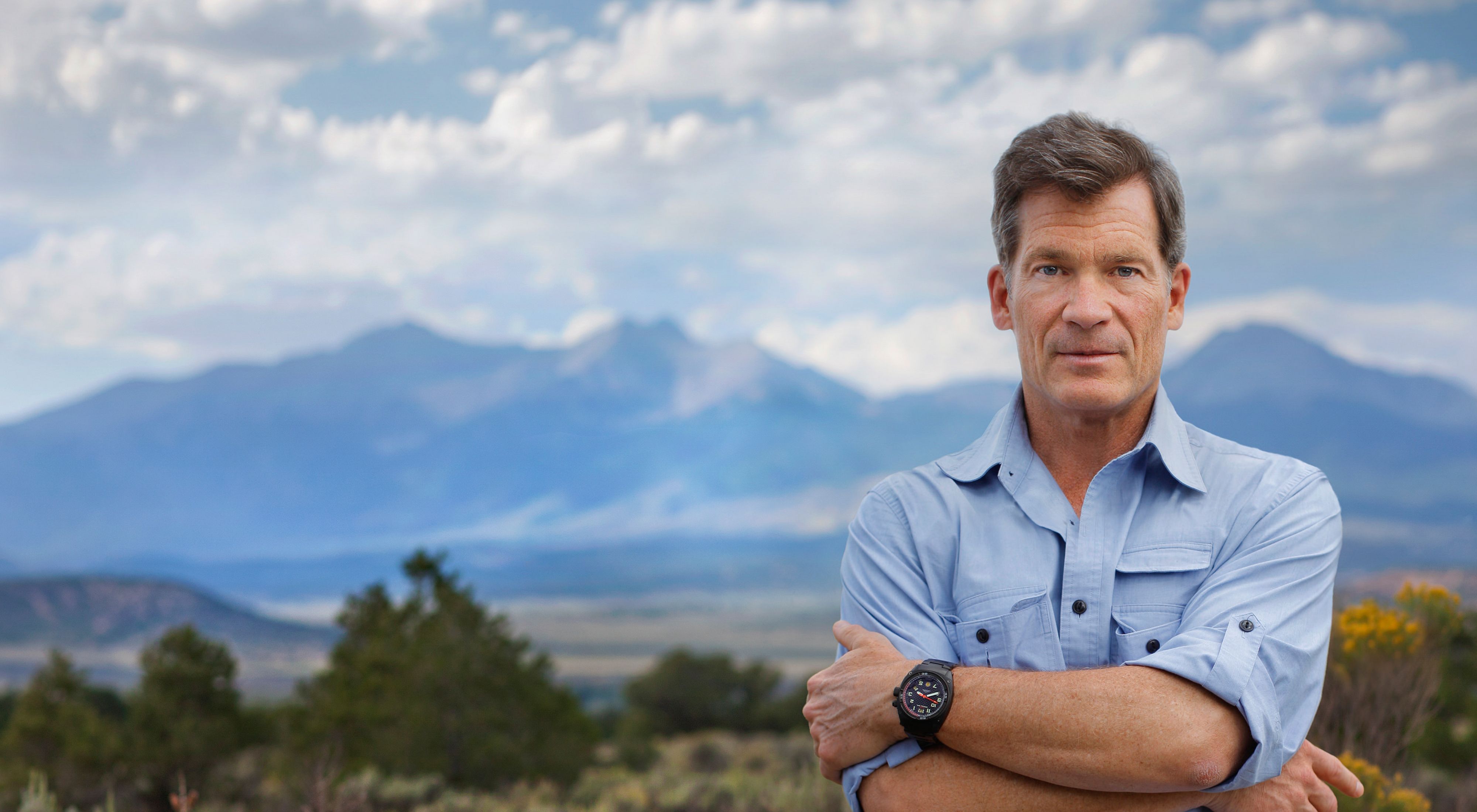 Man in light blue shirt stands with arms folded in front of mountain range.