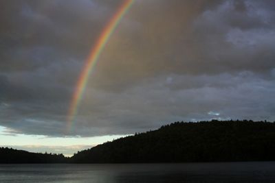 A rainbow breaks through clouds and arches over a lake and forested hill.