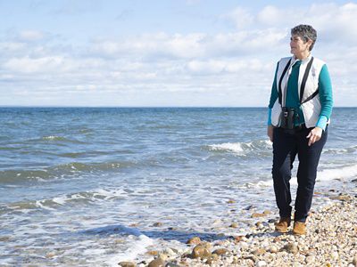 Louise Harrison walking on a shoreline at right looking left to the ocean shore.
