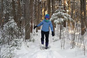 A woman in a blue coat and hat snowshoeing in the woods.