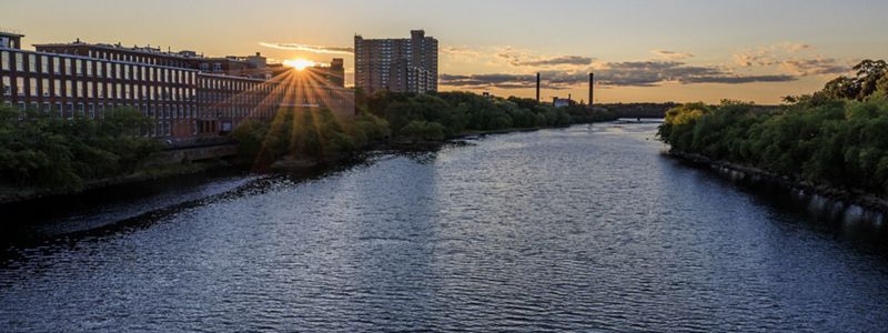 The Merrimack River at dusk lined by old mill buildings on one side and trees on the other.