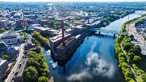 Aerial view of brick mill buildings along the Merrimack River where it passes through the city of Lowell, and branches off into a canal. A bridge extends over the river in the right of the photo.