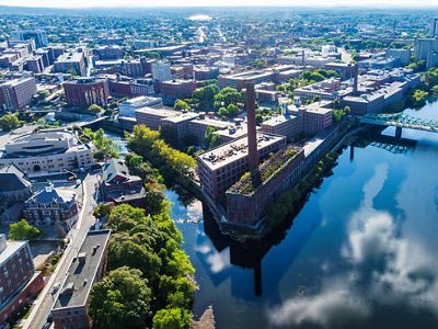 Aerial view of brick mill buildings along the Merrimack River where it passes through the city of Lowell, and branches off into a canal. A bridge extends over the river in the right of the photo.