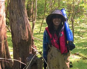 A woman wearing a floppy hat and sunglasses stands in the middle of a swamp.