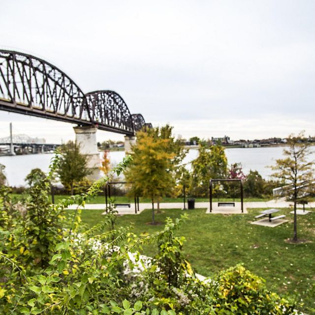 Swings and benches provide places to gather in a public park in Louisville, KY. A railroad bridge rises in the background spanning a wide river.