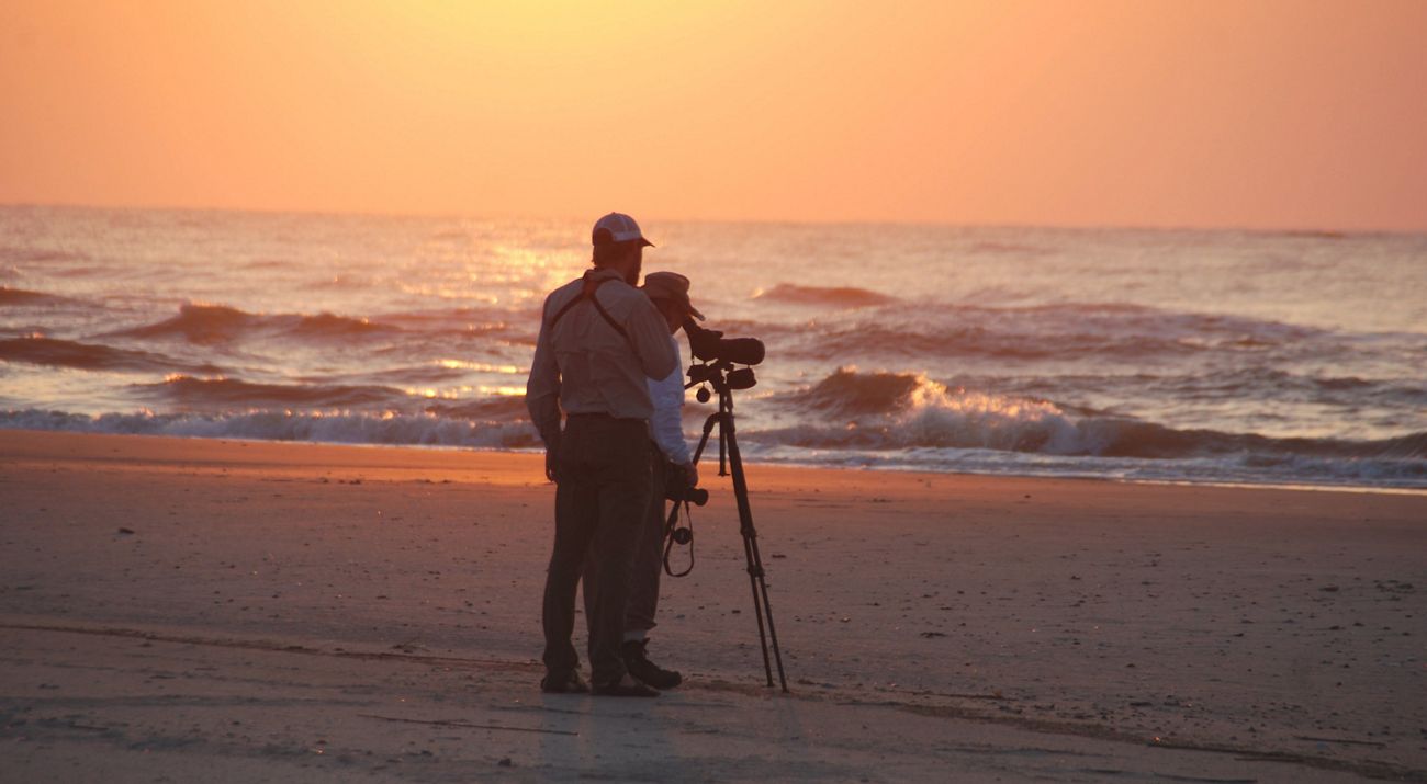 Two people stand on a broad sandy beach, one of them looking through a camera, looking out over ocean waves at sunrise.