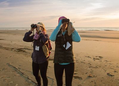 Two people on the shore hold binoculars to their eyes in search of wildlife.