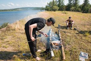 first nation teenage boy roasts marshmallow over fire next to a lake in thaidene nene