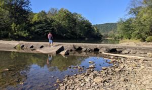 A man stands on an old concrete bridge that has small openings running below it that restrict the flow of water under the bridge. 