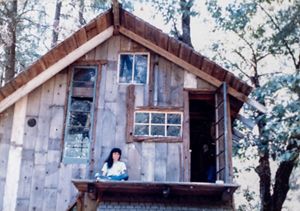 A woman sits on the deck of a wood cabin that is surrounded by trees. 
