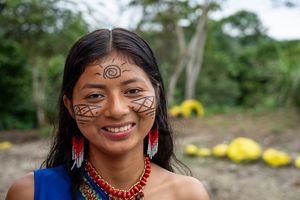 Retrato de mujer joven indígena, con dibujos con pintura negra en su rostro, aros de plumas, collar de cuentas rojo, el pelo oscuro suelto y fondo natural. 