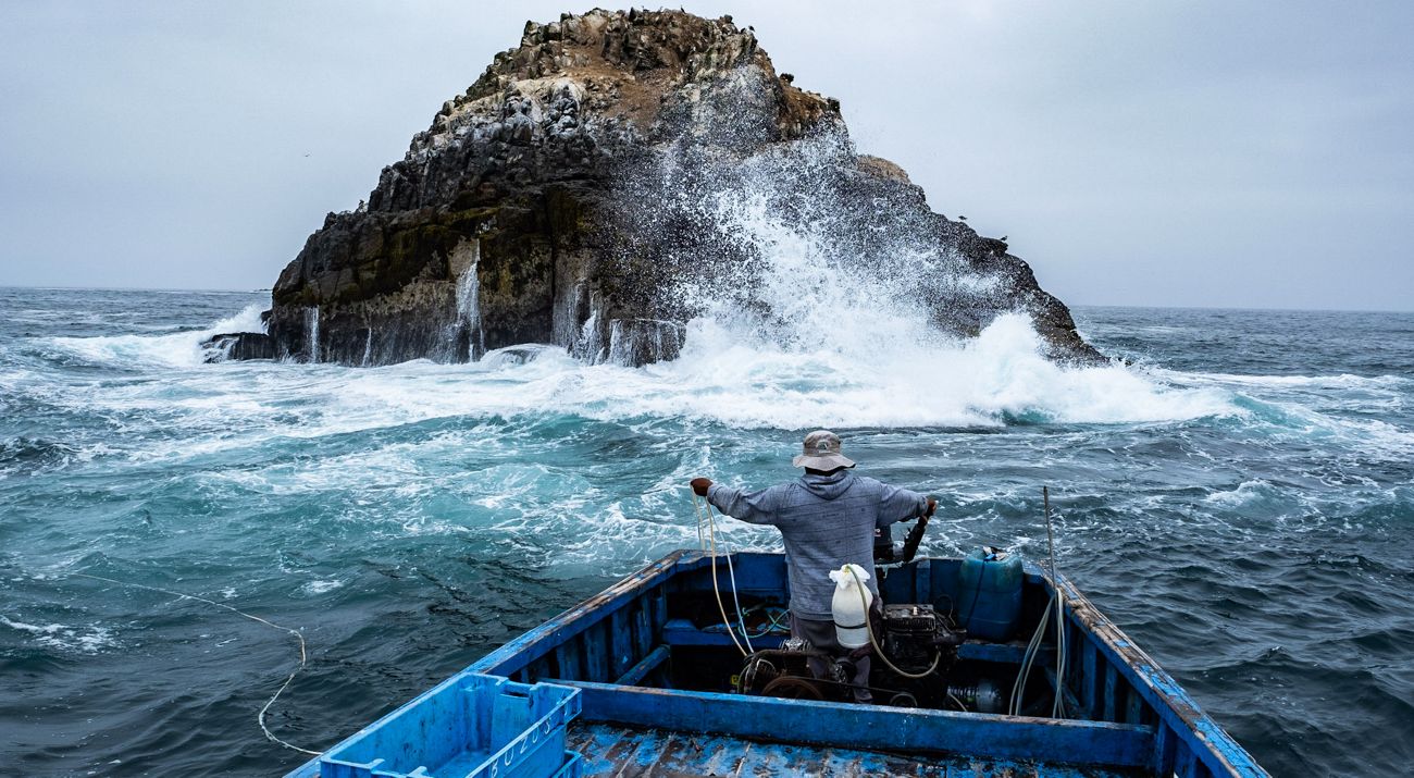 Pescador en un bote de pesca en medio del ocean con una motaña rocosa al frente en Ancón, Perú.