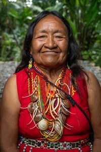 Retrato de mujer indígena madura, vestida de rojo, con collares de conchas, aros amarillos y el pelo oscuro y suelto. 
