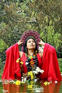 A young indigenous women wearing red traditional clothes and artisanal jewelry, submerged in the water to her waist, surrounded with flowers on the water and trees behind her. 