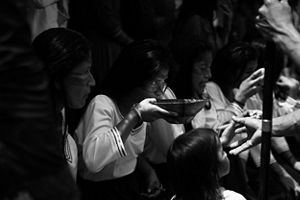 A black and white picture showing a group of indigenous woman, wearing white clothes, on a ceremony, where one of them is drinking something from a bowl. 