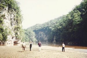 Grupo de personas caminando por la ribera de un río, rodeado de un cañón con vegetación, foto antigua de archivo. 