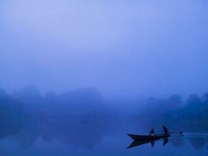 A silhouette of a canoe with two people on board navigating on a river at dawn, with predominating blue tones and mist. 