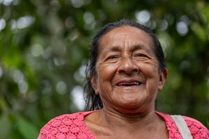 A woman smiles as she stands in front of lush green trees, radiating joy and warmth in a natural setting.