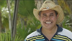 Un hombre con un sombrero de paja sonríe cálidamente a la cámara, mostrando una actitud amigable.