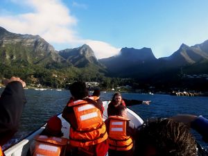 personas del espaldas con chaleco salvavidas en un bote con montañas en el fondo.