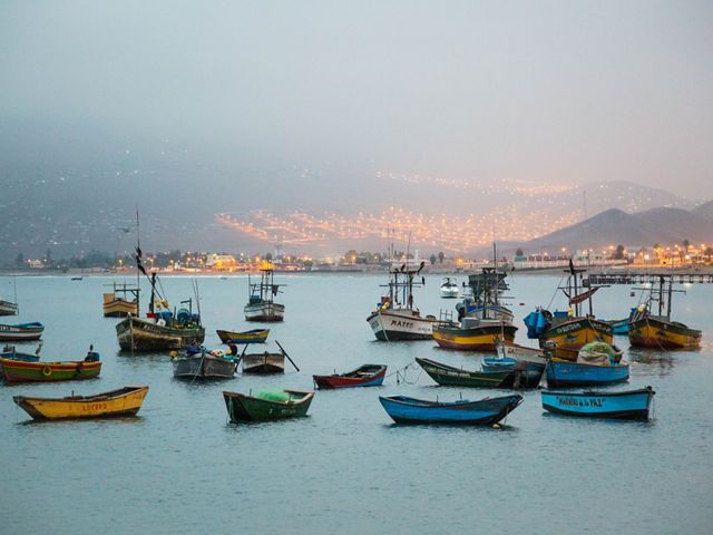 Small fishing boats float in the foreground near a dusky, electrified coastal mountain village