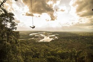 Persona suspendida en tirolesa sobre un paisaje de bosque, copas de arboles y rios.