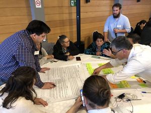 Un grupo de personas participa en un taller colaborativo alrededor de una mesa, discutiendo sobre una hoja grande con texto. El ambiente es concentrado e interactivo.
