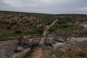 Vista aérea de varios vehículos cruzando un estrecho puente de madera sobre un río rocoso en un paisaje accidentado.