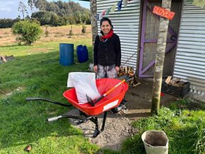  Una persona sonríe junto a una carretilla roja llena de herramientas, cerca de un pequeño cobertizo de chapa ondulada, en medio de un paisaje rural.