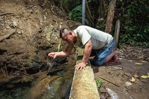 Un hombre se arrodilla junto a un arroyo, ahuecando las manos para beber agua de la fuente que fluye.