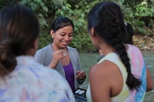 Una mujer sonriente está de pie junto a un grupo de personas, todas aparentemente participativas y felices en un ambiente social.